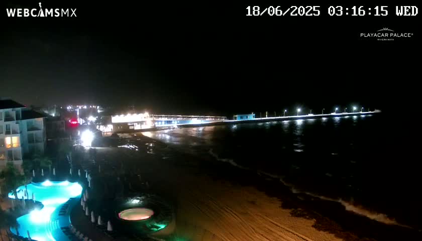 A nighttime view of a resort area shows a lit-up pier extending into a dark ocean with gentle waves lapping the shore, and a pool area with lit-up features is visible in the foreground.