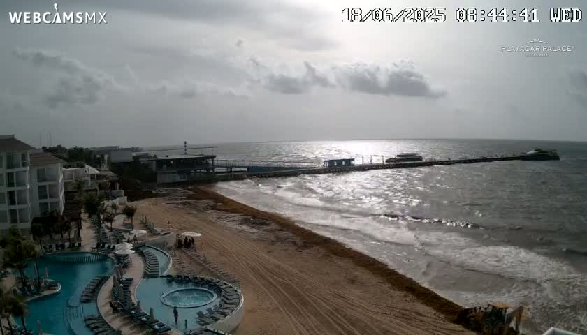 An overcast day shows a beach with a resort, pool, and pier extending into a wavy ocean.