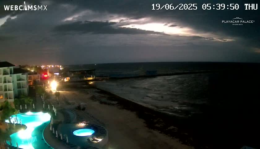 A resort with illuminated pools sits on a beach at night under a dark, cloudy sky with hints of a break in the clouds showing a lighter sky.