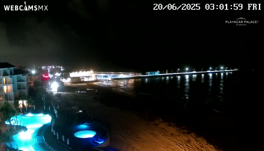 A nighttime view of a beach at a resort with a lit pier extending into the dark water, and illuminated pools near a building.