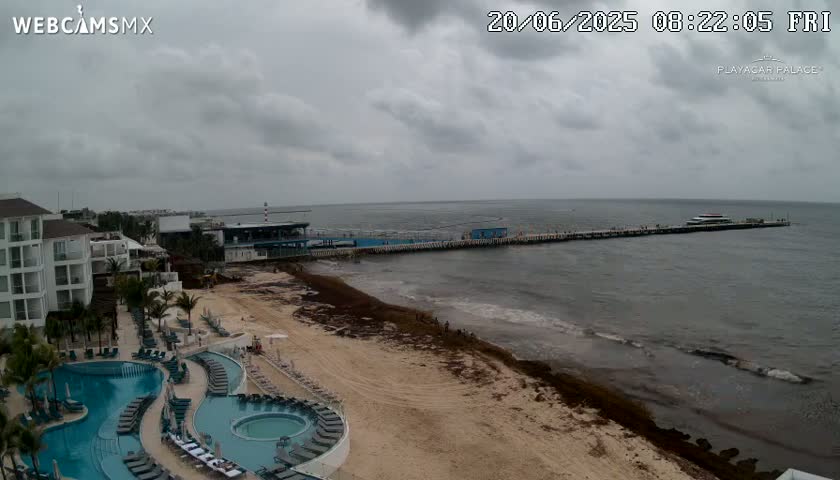 A resort with multiple pools overlooks a sandy beach and a long pier extending into a gray, cloudy ocean.