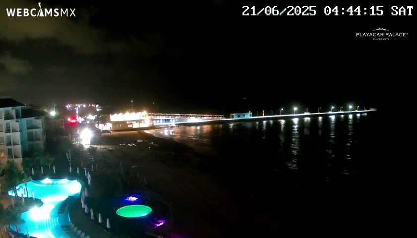 A nighttime view of a resort with illuminated pools, alongside a brightly lit pier extending into dark water under a partly cloudy sky.