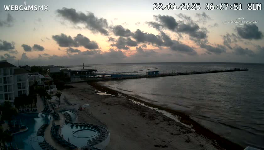 A sunrise over a calm ocean, showing a beach with a resort, pool, and pier.
