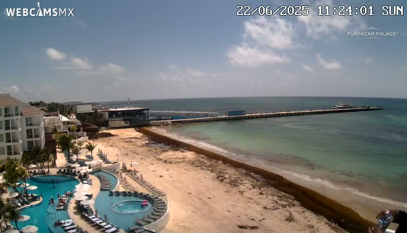 A resort with multiple pools overlooks a sandy beach with seaweed and a long pier extending into a calm, sunny ocean.