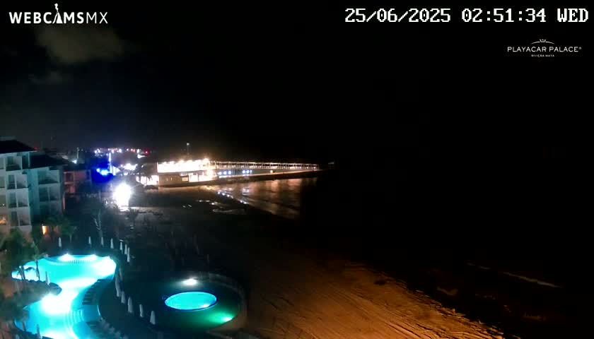 A nighttime view of a beach with a resort featuring illuminated pools and a pier extending into the dark ocean.