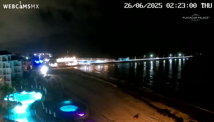 A nighttime view of a beach with a brightly lit pier extending into the calm water, next to a resort with illuminated pools.
