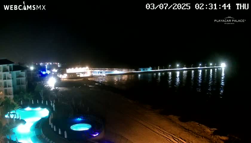 A brightly lit pier extends into dark water at night next to a resort with illuminated pools.