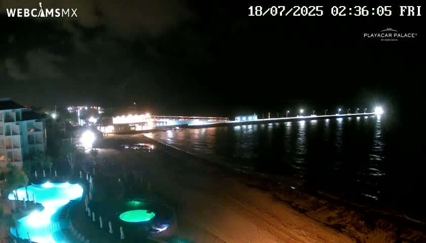 A nighttime view of a beach resort with illuminated pools, a long pier extending into the dark ocean under a partly cloudy sky.