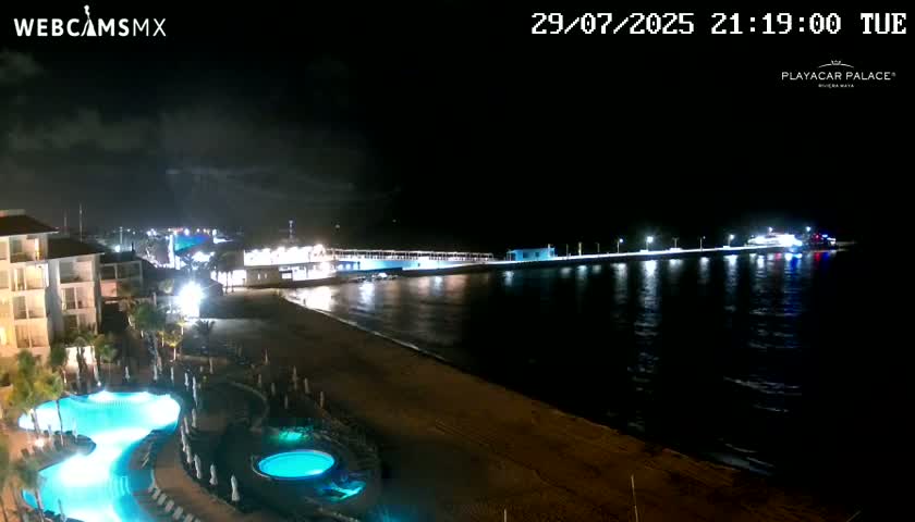 A nighttime view of a beach resort with illuminated pools, a long pier extending into the dark ocean under a partly cloudy sky.