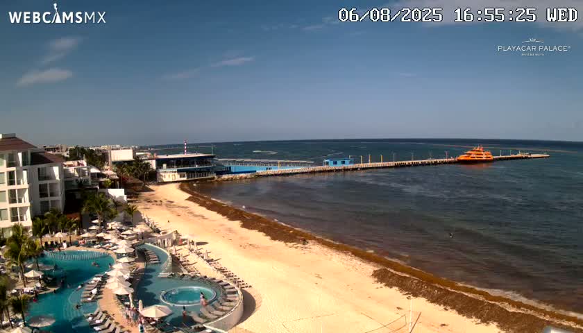 An aerial view shows a beach with seaweed along the shoreline, a resort with multiple pools, and a pier with a docked orange ferry, all under a clear, sunny sky.