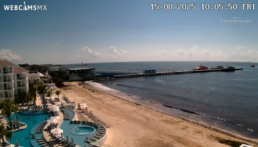 An aerial view shows a beach with seaweed along the shoreline, a resort with multiple pools, and a pier with a docked orange ferry, all under a clear, sunny sky.