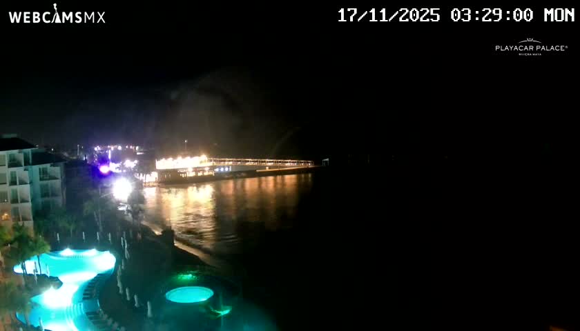 A nighttime view reveals a brightly lit resort with glowing blue swimming pools bordering a calm ocean, where a long illuminated pier extends under a clear, dark sky.