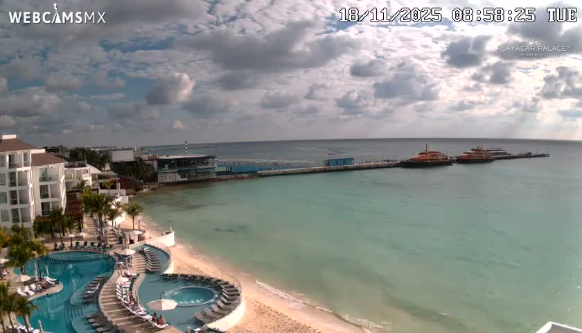 A nighttime view reveals a brightly lit resort with glowing blue swimming pools bordering a calm ocean, where a long illuminated pier extends under a clear, dark sky.