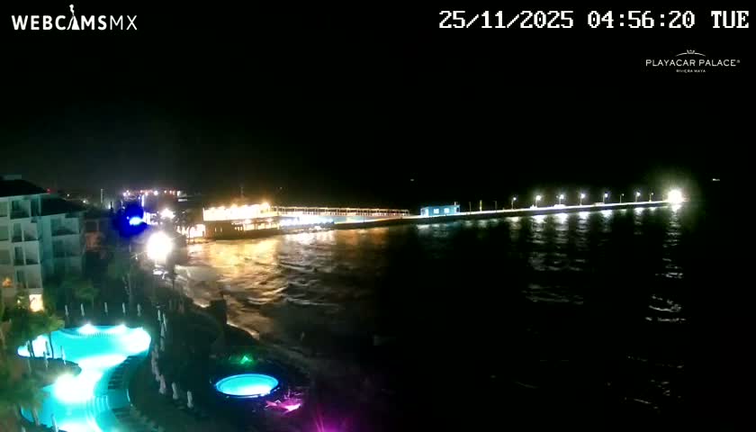 A brightly lit resort complex with glowing turquoise swimming pools lines the shore of an ocean at night, where a long, illuminated pier extends into the clear, dark expanse.