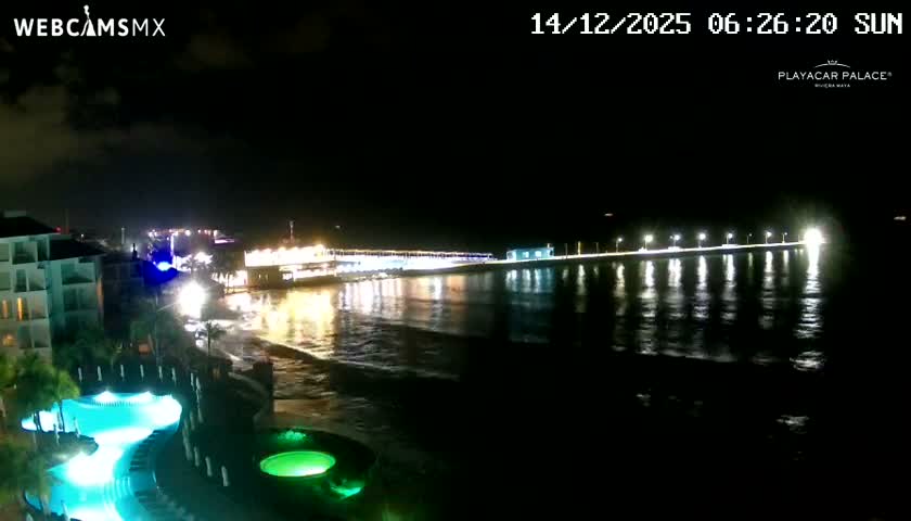 A brightly lit resort complex with glowing turquoise swimming pools lines the shore of an ocean at night, where a long, illuminated pier extends into the clear, dark expanse.