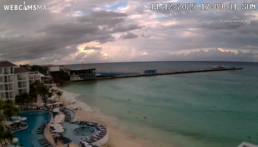 A brightly lit resort complex with glowing turquoise swimming pools lines the shore of an ocean at night, where a long, illuminated pier extends into the clear, dark expanse.