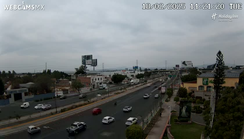 A multi-lane highway with moderate traffic is seen on an overcast day, surrounded by buildings and landscaping.