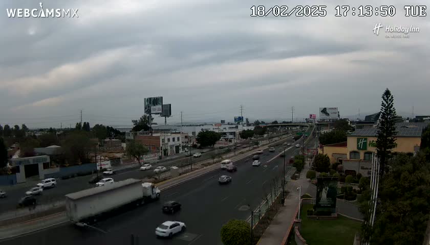 A mostly cloudy day reveals a highway scene with moderate traffic, featuring buildings, and greenery alongside the road.