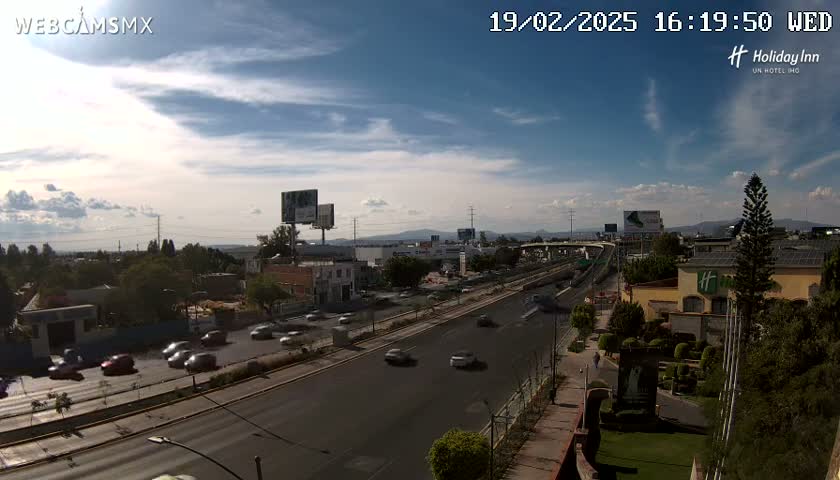 A partly cloudy day reveals a highway with moderate traffic, surrounded by buildings and vegetation, including a Holiday Inn.