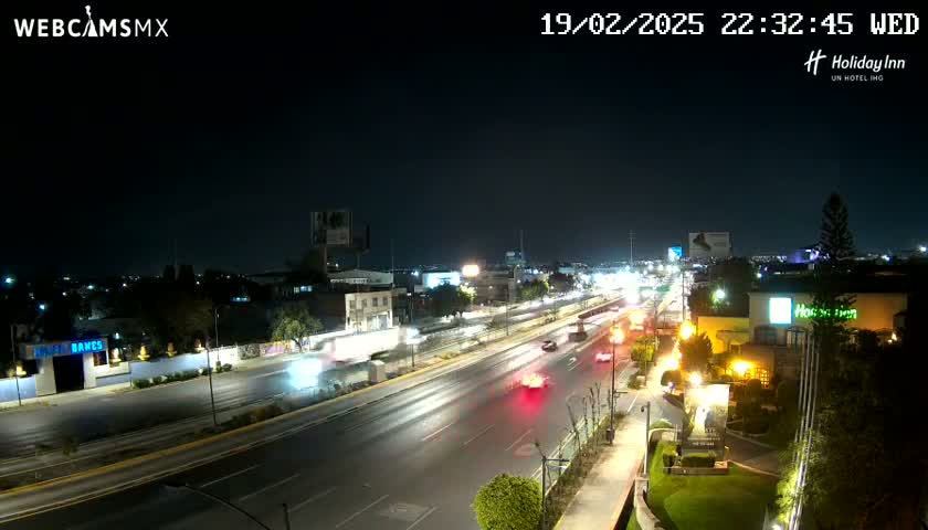 A nighttime, city highway scene shows several vehicles driving on a multi-lane road, passing buildings and businesses under a clear sky.