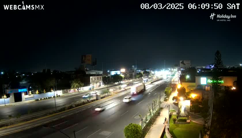 A nighttime, high-angle view of a multi-lane road with sparse traffic, surrounded by buildings and some vegetation.