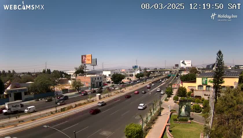 A sunny day reveals a multi-lane highway with moderate traffic, alongside buildings and green landscaping.