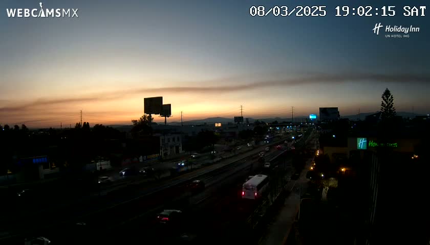 An elevated view of a highway at sunset, with traffic moving along multiple lanes, billboards visible in the distance, and buildings and trees lining the roadside.