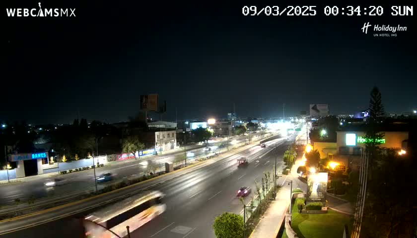 A nighttime view of a multi-lane highway with sparse traffic, surrounded by buildings and landscaping under clear skies.