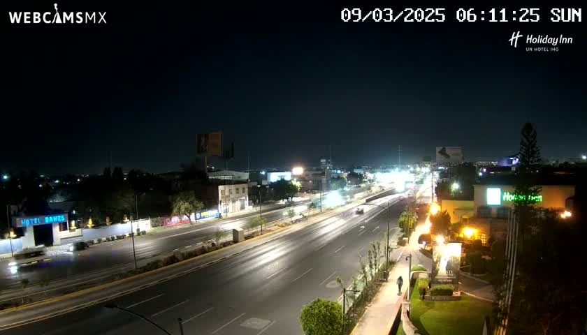 A nighttime view of a multi-lane highway with sparse traffic, flanked by buildings and landscaping under clear skies.