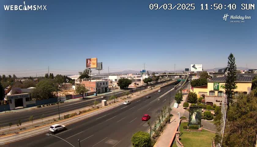 A sunny day reveals a multi-lane highway with sparse traffic, flanked by buildings, landscaping, and billboards, under a clear blue sky.