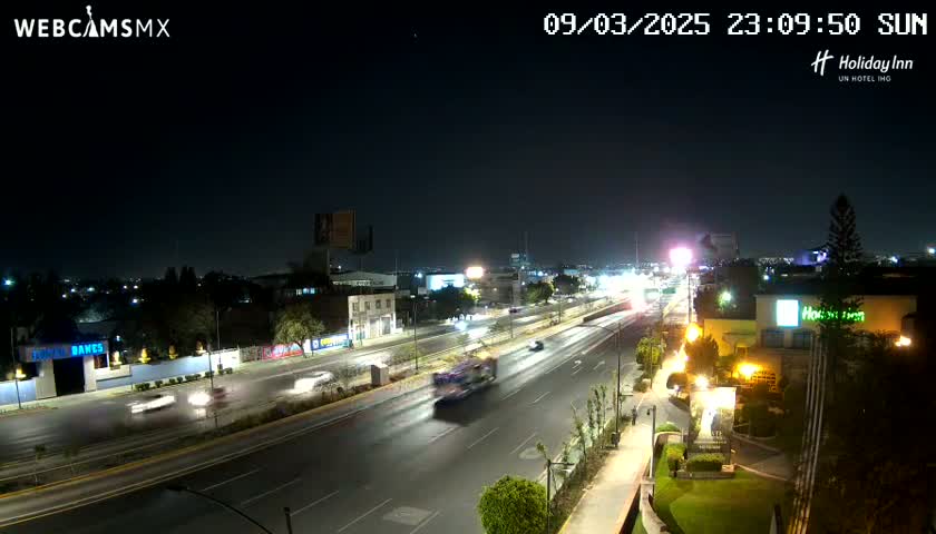 A nighttime, slightly overcast view of a multi-lane highway with sparse traffic, surrounded by buildings and illuminated by streetlights.