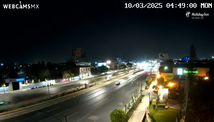 A nighttime view of a multi-lane highway with sparse traffic, surrounded by buildings and landscaping under a clear sky.