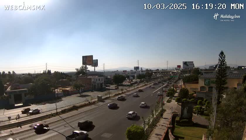 A sunny day reveals a multi-lane highway with moderate traffic, surrounded by buildings, landscaping, and billboards, in a suburban or urban area.