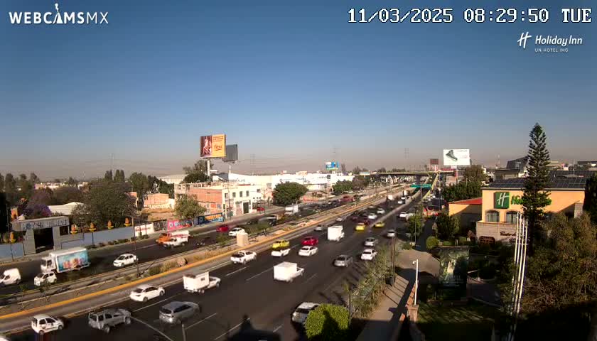 A highway with moderate traffic is seen on a clear, sunny day, with buildings and greenery lining the roadside.
