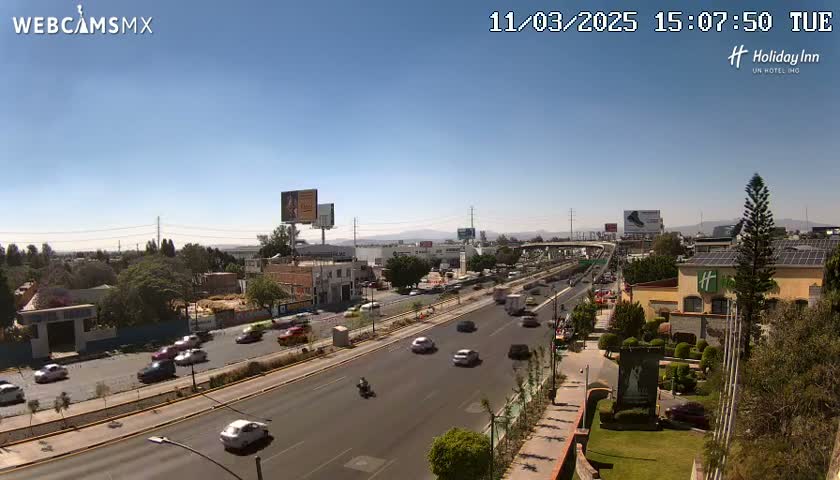 A sunny day reveals a highway scene with several cars driving, buildings lining the road, and green landscaping visible.