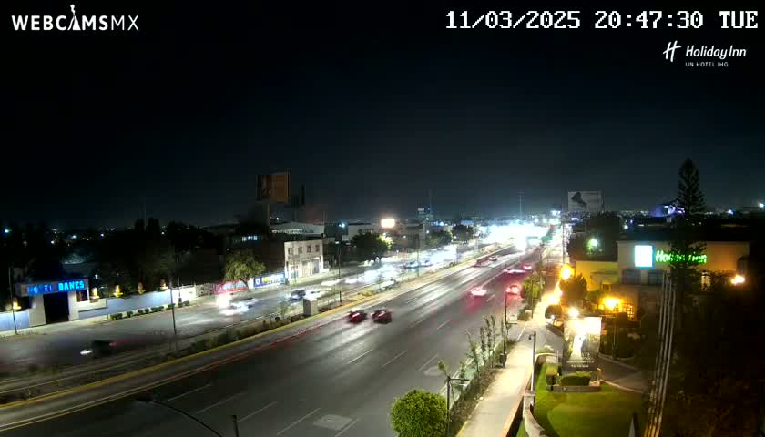 A nighttime, high-angle view of a multi-lane highway with moderate traffic, surrounded by buildings and some vegetation under a clear sky.
