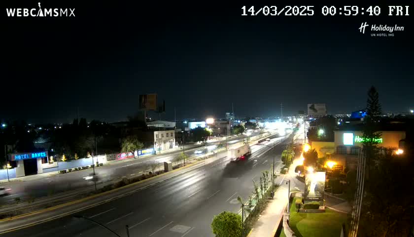 A nighttime, high-angle view shows a multi-lane highway with sparse traffic, lined with buildings and illuminated by streetlights.