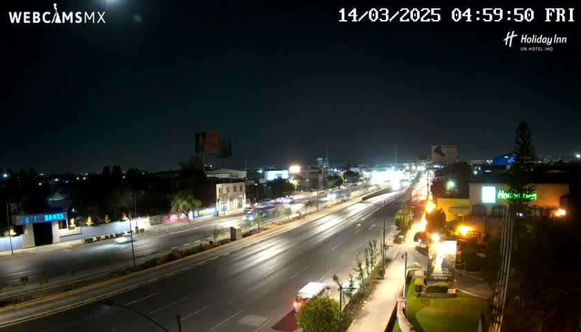 A nighttime, slightly moonlit view of a multi-lane highway with sparse traffic, flanked by buildings and landscaping.