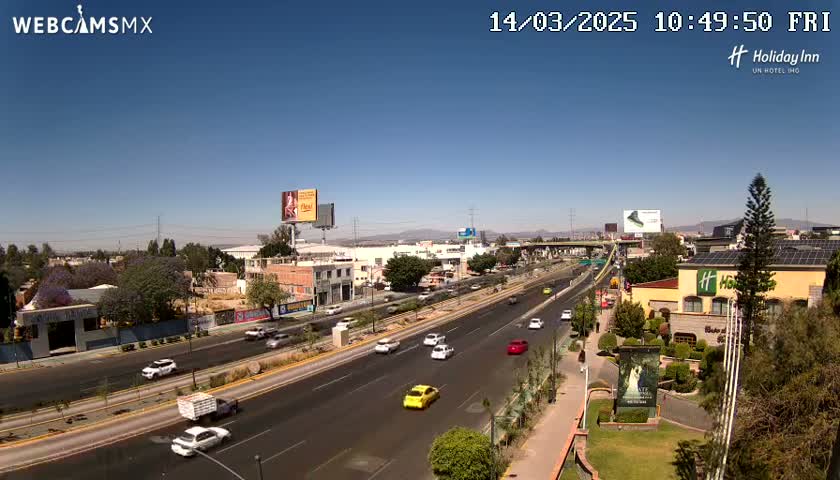 A sunny day reveals a multi-lane highway with light traffic, flanked by buildings, vegetation, and billboards, under a clear blue sky.