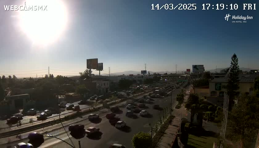 A sunny day reveals heavy traffic on a multi-lane highway, surrounded by buildings and greenery.