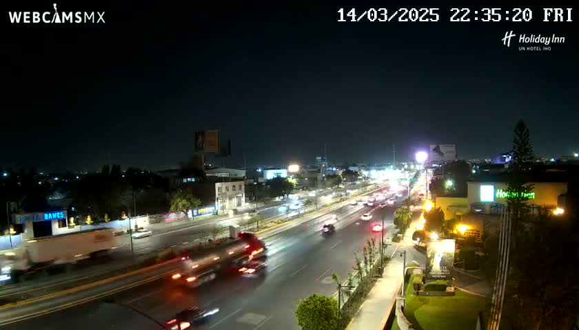 A nighttime, high-angle view of a multi-lane highway with moderate traffic, surrounded by buildings and trees under clear skies.