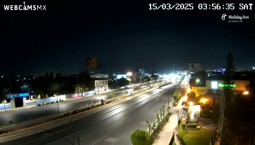 A nighttime view of a multi-lane highway with sparse traffic, surrounded by buildings and streetlights under a clear sky.