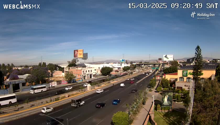 A highway with moderate traffic is seen on a sunny day, with buildings and landscaping visible on either side.