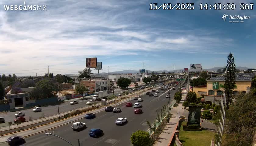 A partly cloudy day shows a multi-lane highway with moderate traffic, flanked by buildings, landscaping, and billboards.