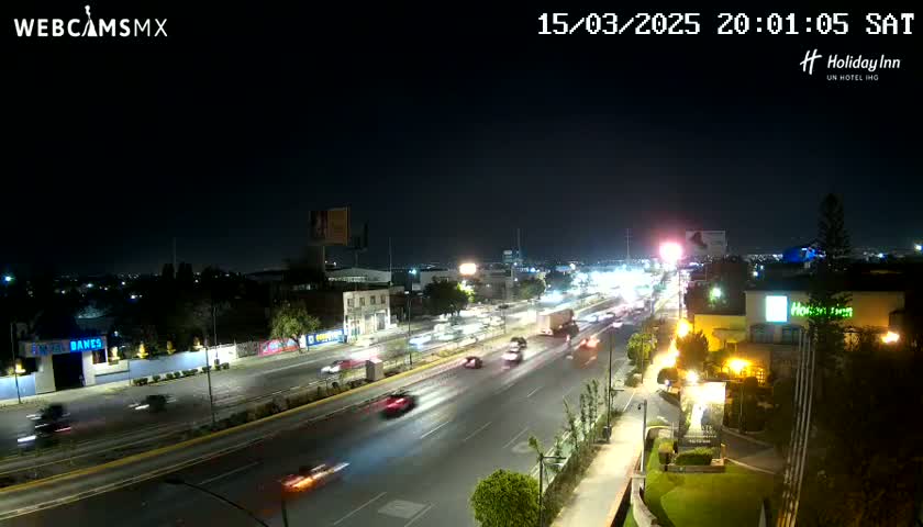 A nighttime, high-angle view of a multi-lane highway with moderate traffic, surrounded by city buildings and vegetation.