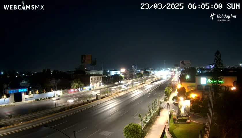 A nighttime, brightly lit highway scene with sparse traffic, surrounded by buildings and landscaping.
