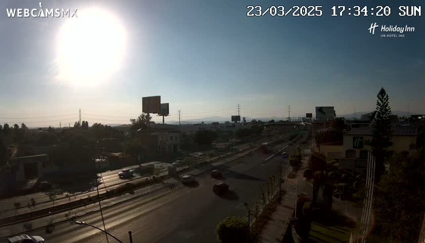 A sunny day reveals a highway scene with several vehicles, buildings, and billboards, all under a bright, mostly clear sky.