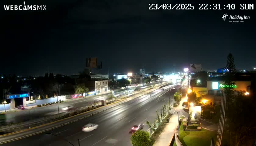 A nighttime, high-angle view shows a multi-lane highway with cars driving on it, surrounded by buildings and trees under a clear sky.