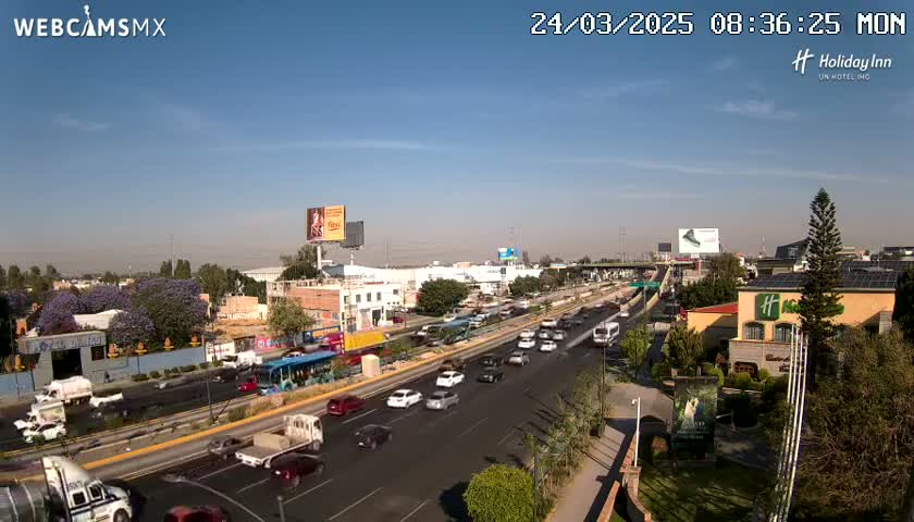 A highway with moderate traffic under a clear blue sky, surrounded by buildings and vegetation.