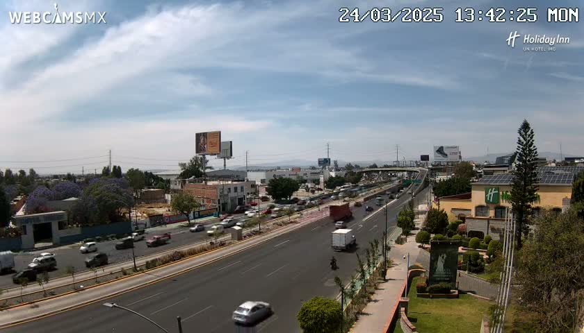 A partly cloudy day reveals a multi-lane highway with moderate traffic, bordered by buildings, landscaping, and a body of water in the distance.